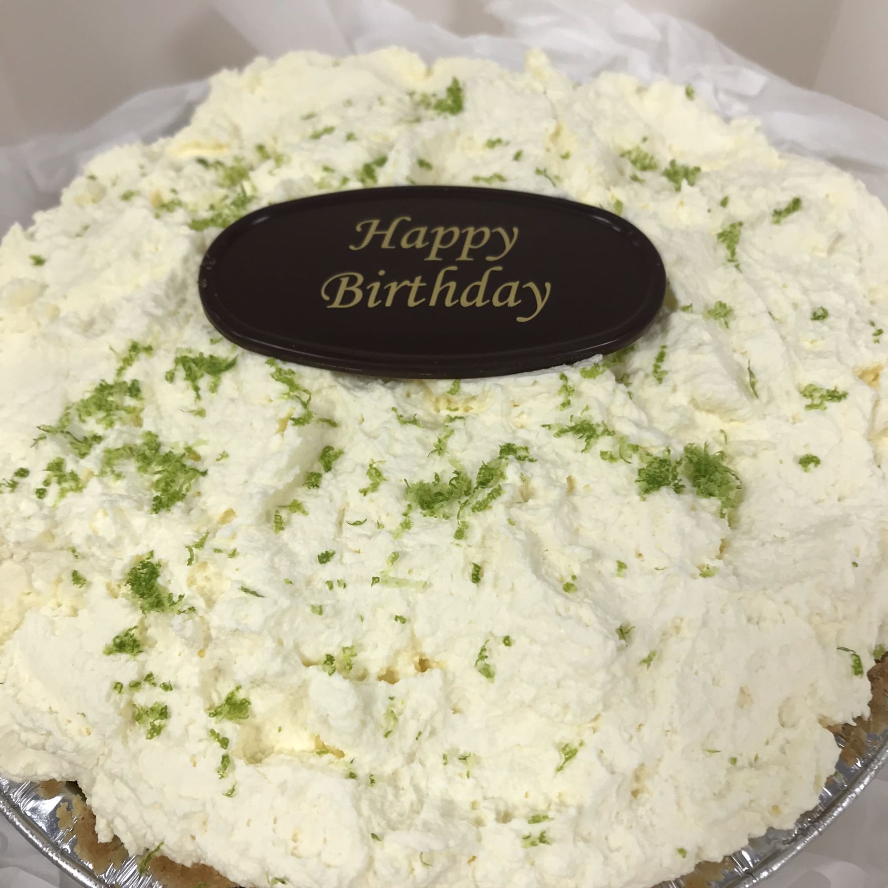 A cake topped with fluffy white cream and sprinkled with green zest, featuring a black oval chocolate plaque in the center that says 'Happy Birthday' in gold letters. Infused with tropical flavor and authentic Key West key limes, this treat is ready for a birthday celebration atop a white surface.